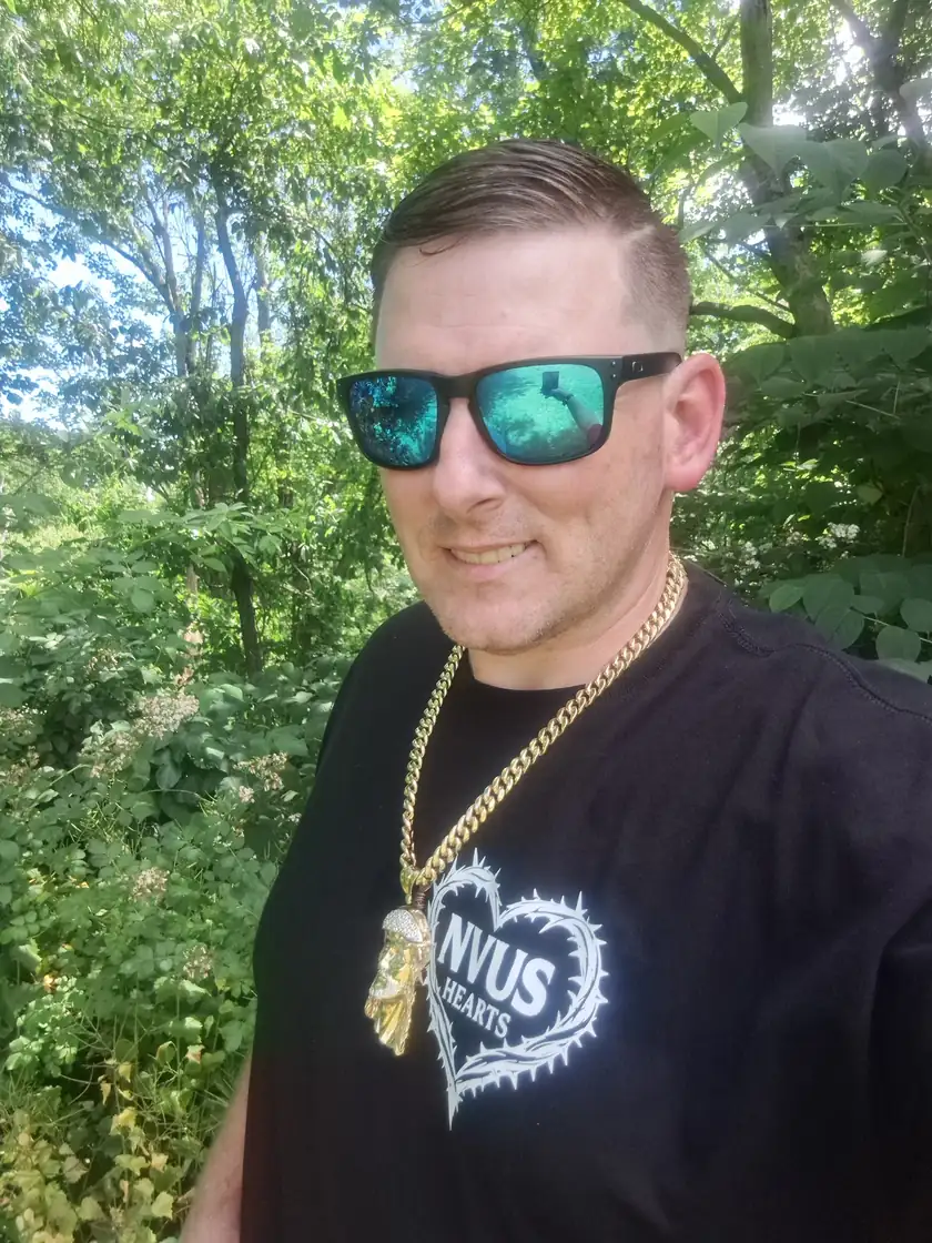 Keith, founder of NVUS Hearts, outdoors among green trees, wearing sunglasses, a gold chain and the black NVUS Hearts tee with the thorn-heart logo