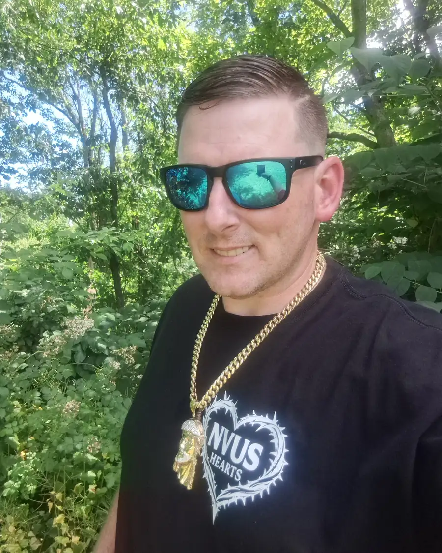 Keith, founder of NVUS Hearts, outdoors among green trees, wearing sunglasses, a gold chain and the black NVUS Hearts tee with the thorn-heart logo